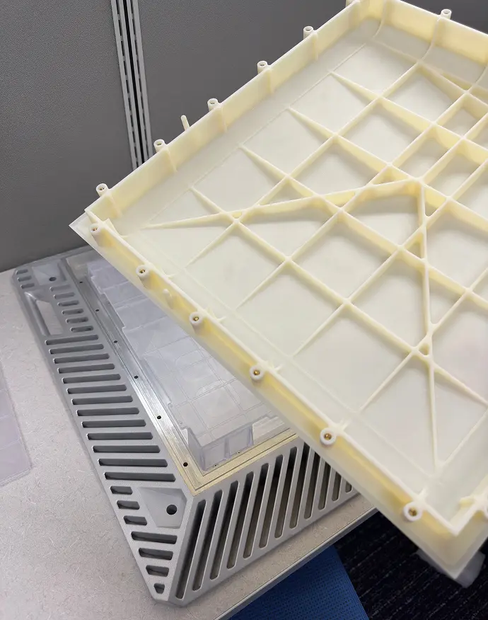 Close-up of a cream-colored plastic tray with grid and support ribs being held above a gray vented plastic base on a beige surface.