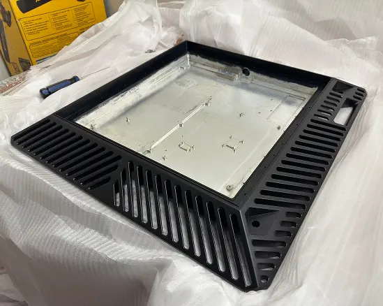 A black metal grate with parallel slats on three sides, surrounding a metallic square panel with screw holes and fixtures, placed on a white textured surface.