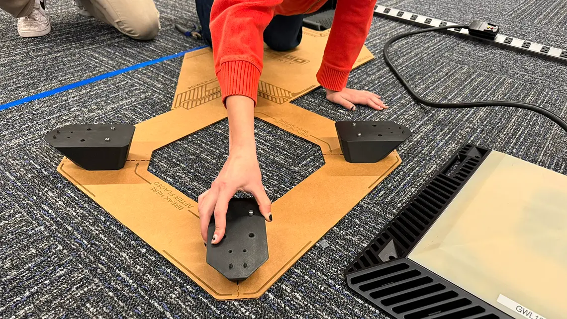 Person kneeling on carpeted floor assembling or positioning black plastic components on a cardboard template with blue tape lines and measuring tape in the background.