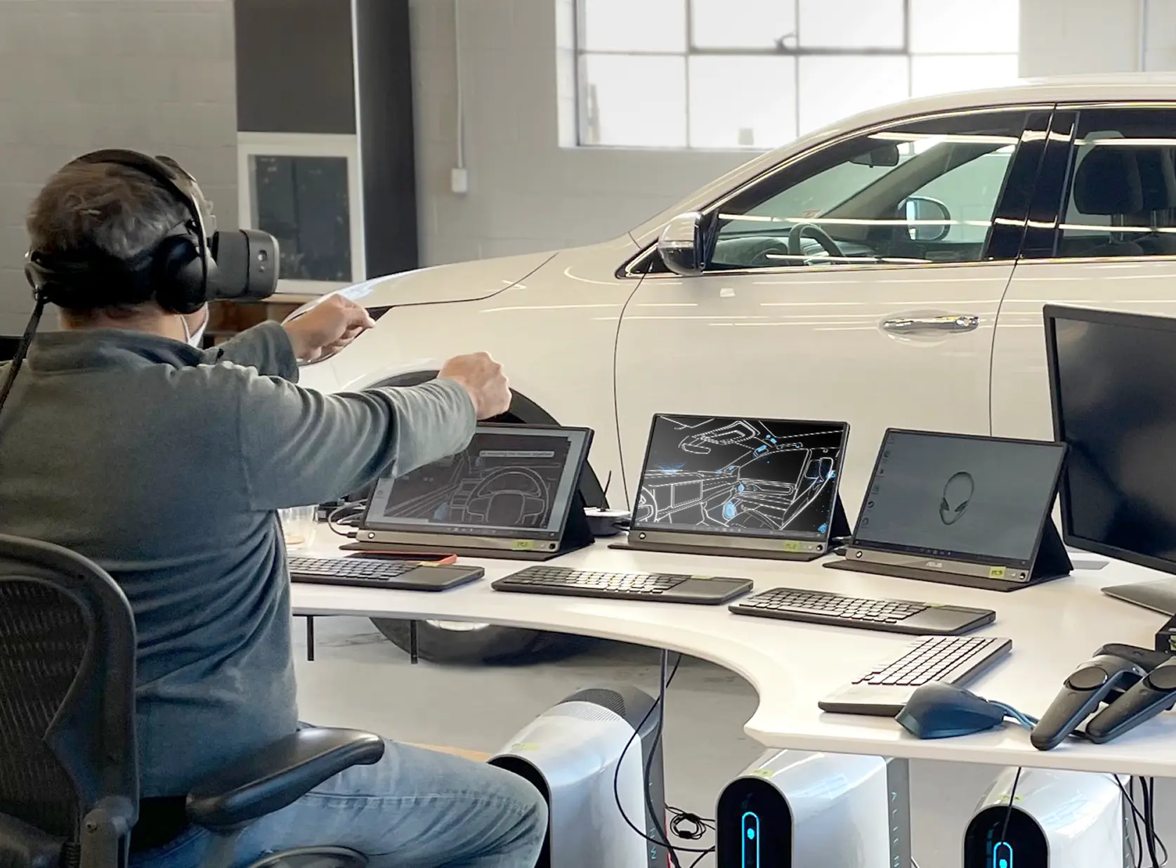 Person wearing a virtual reality headset and headphones sitting at a curved desk with three tablets displaying car interior design images and multiple keyboards, mice, and VR controllers, with a white car in the background inside a modern workspace.