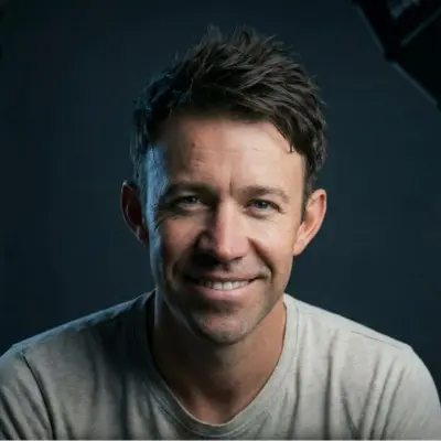 Portrait of a smiling man with short, tousled dark hair, wearing a light gray shirt against a dark background.