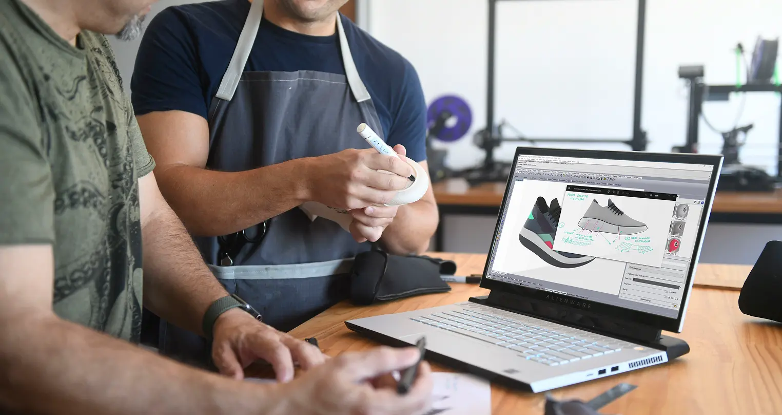 Two men collaborating on a shoe design at a wooden table; one man in a gray apron holds a white sneaker and marker, while the other gestures with a pen. A laptop displays 3D shoe models with design notes and color options.