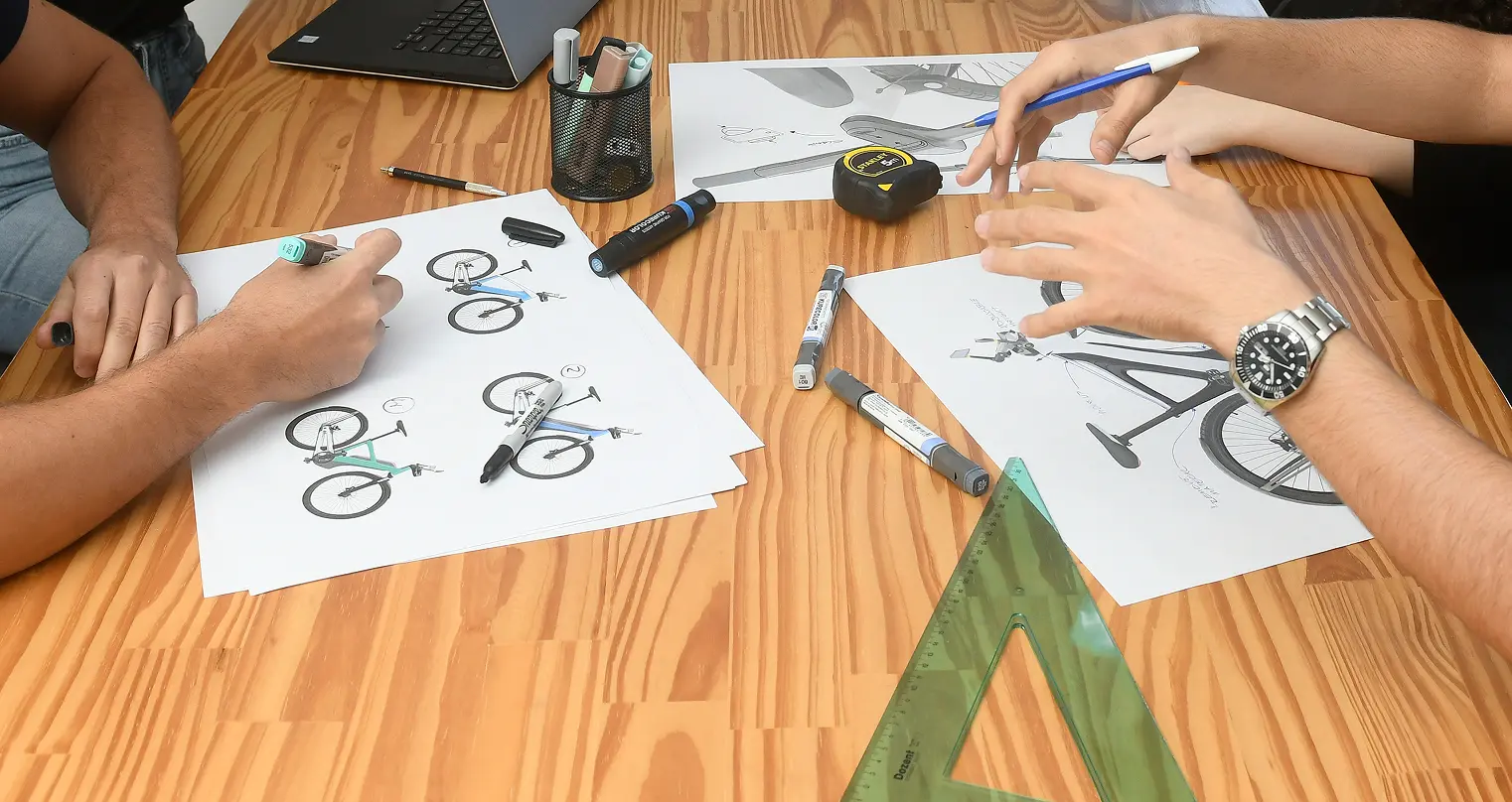 Two people at a wooden table working on bicycle design sketches, using pens, markers, a ruler, and a tape measure, with a laptop nearby.