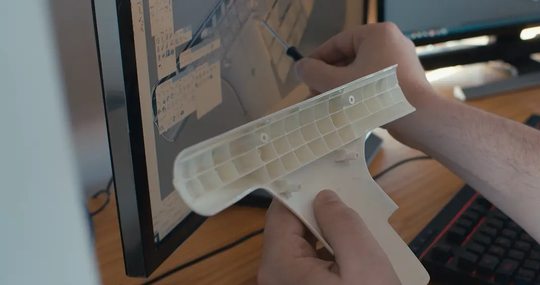 Person holding a white plastic part and using a screwdriver to work on a computer screen displaying technical drawings or a CAD design, with a keyboard and another monitor in the background.