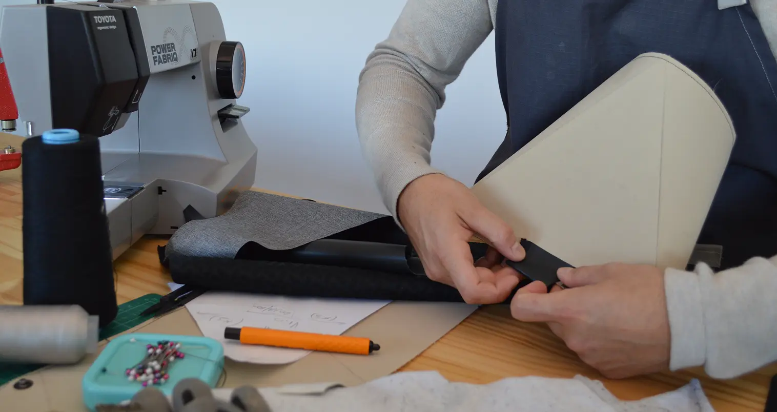 Person working on upholstery, attaching black fabric or material to a beige cushion or chair part, with a sewing machine, scissors, pins, and pen on a wooden work surface.