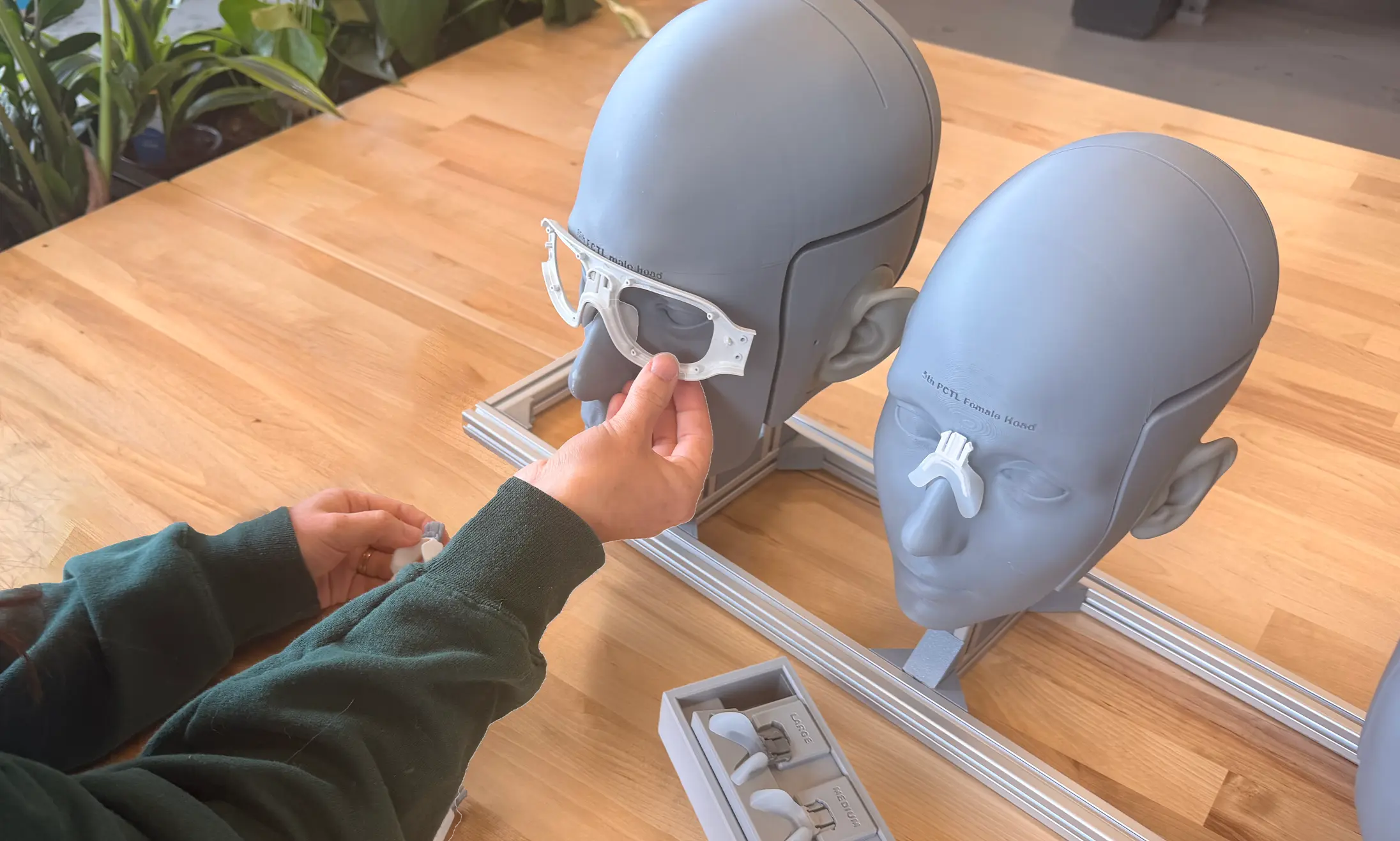 Two gray mannequin heads on a wooden table, one labeled '5th PCYL male head' with a white frame-like object held in front of it, and the other labeled '5th PCYL female head' with a white nose bridge attached; a person wearing a green long sleeve is holding additional white components near a box labeled with sizes.