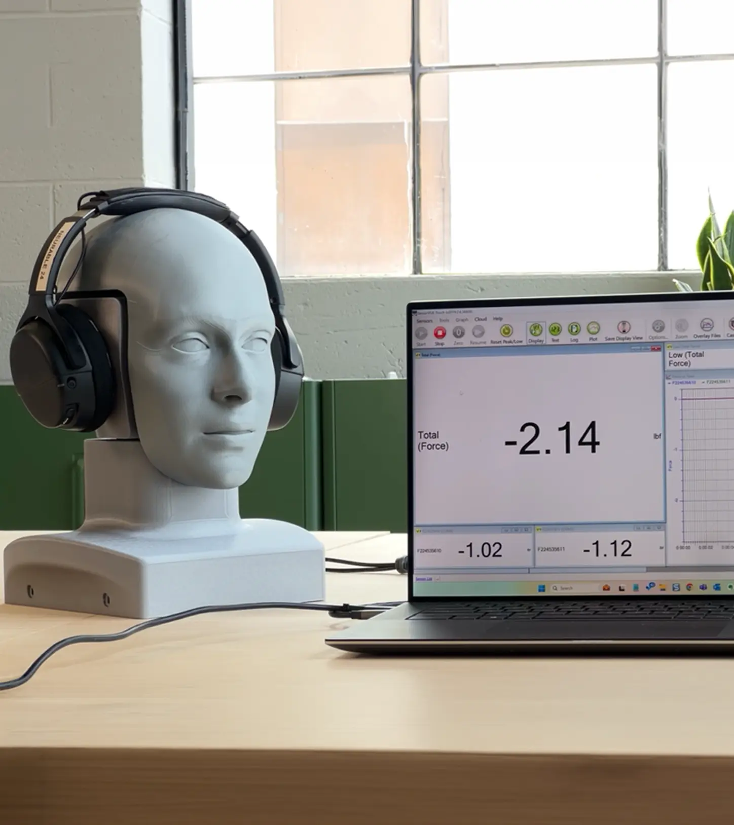 White mannequin head wearing black headphones on a table next to a laptop displaying force measurement software with values including a total force of -2.14.
