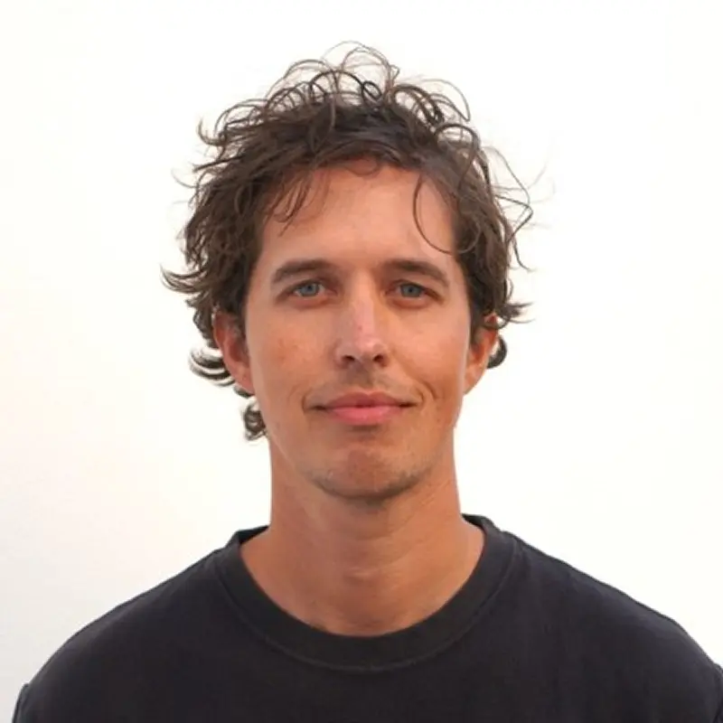 Man with curly brown hair and blue eyes wearing a black crew neck shirt, smiling slightly against a plain white background.