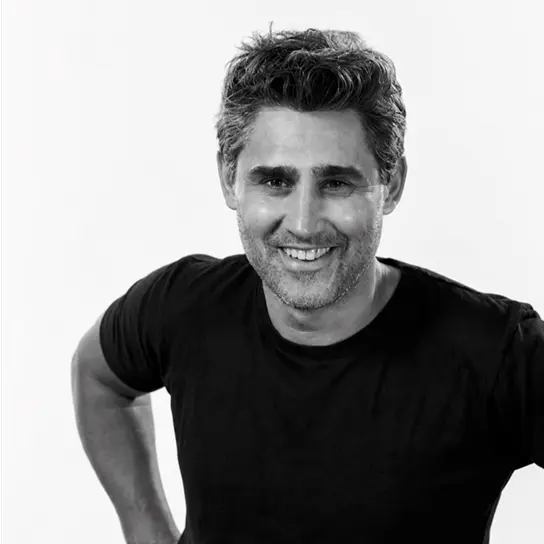 Smiling man with short dark hair and slight stubble wearing a black t-shirt, posing with hands on hips against a plain light background.