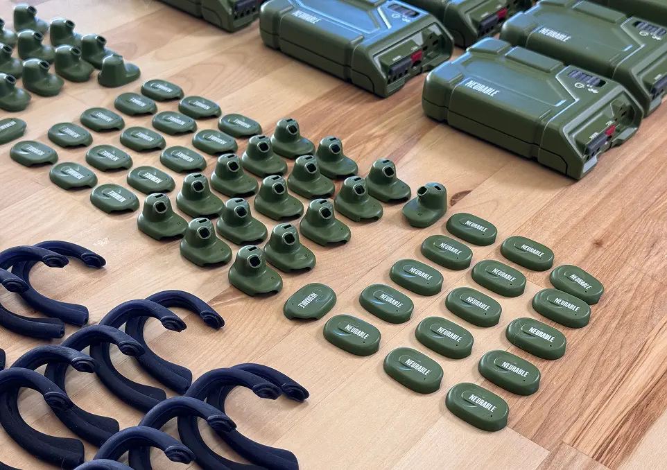Several rows of green electronic devices and components neatly arranged on a wooden surface, including small units labeled 'NEURABLE', circular holders, and black curved plastic parts.