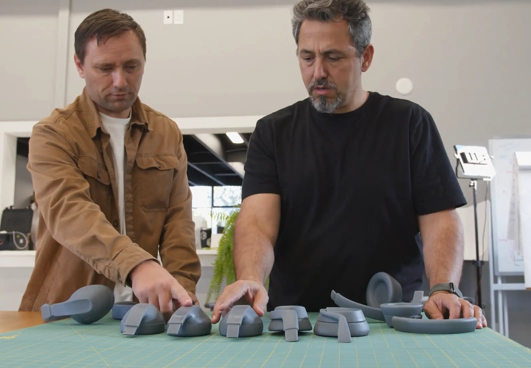 Two men examine and arrange various gray headphone ear pad and headband components on a green cutting mat on a table in a well-lit workspace.