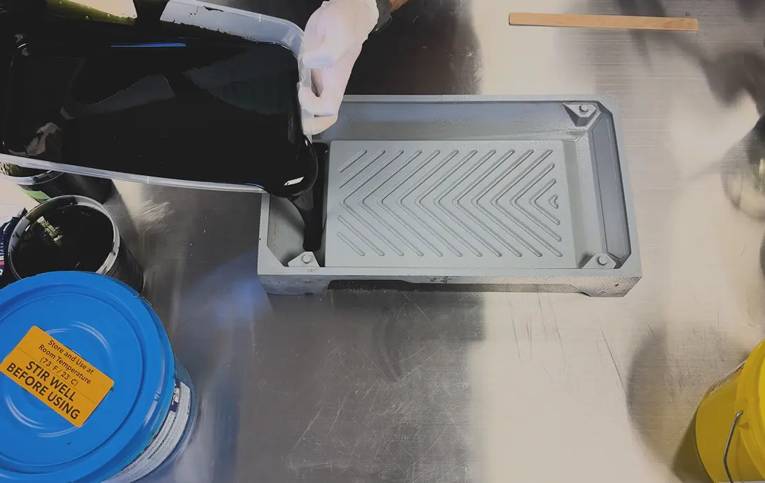 Person wearing a white glove pouring black liquid from a container into a rectangular gray mold tray with a textured surface on a metallic table, next to cans and a yellow bucket.