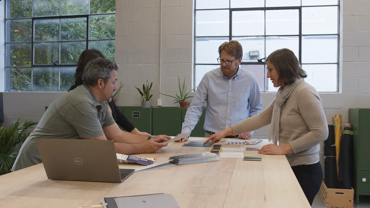 Four people standing around a wooden table in a bright office, discussing color swatches and materials, with a laptop and guidebook on the table.