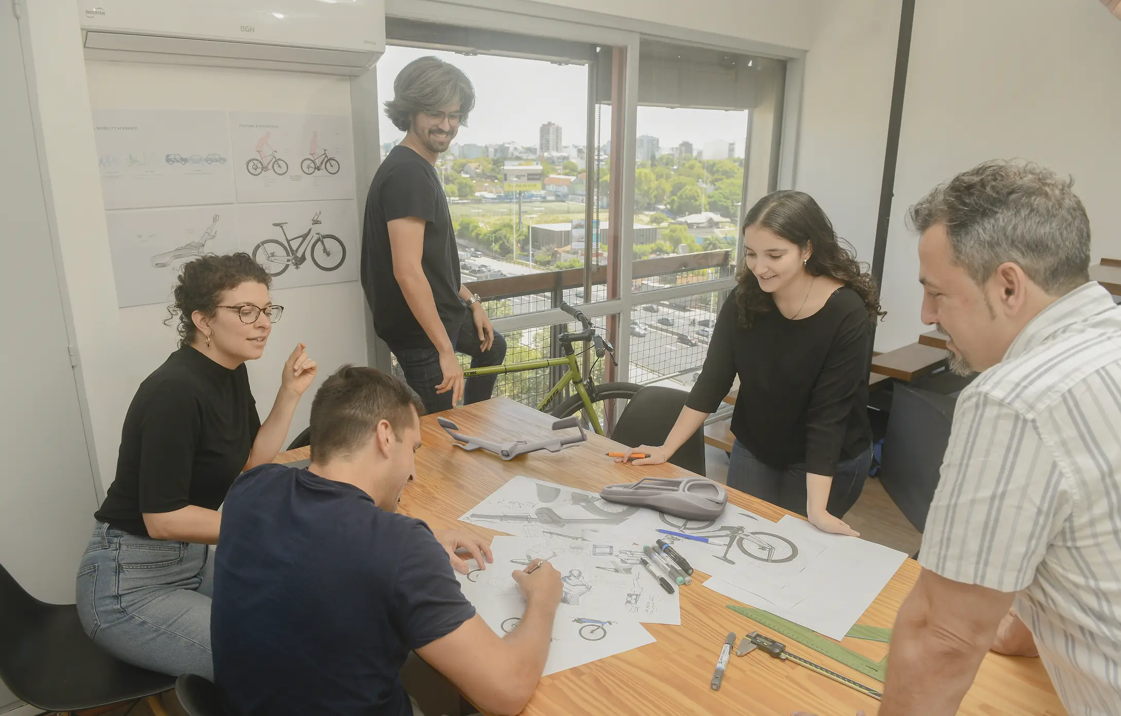 Five designers collaborating around a table with bicycle design sketches, 3D-printed e-bike parts, markers, and measuring tools, with a yellow bike and cityscape visible through a large window behind them.