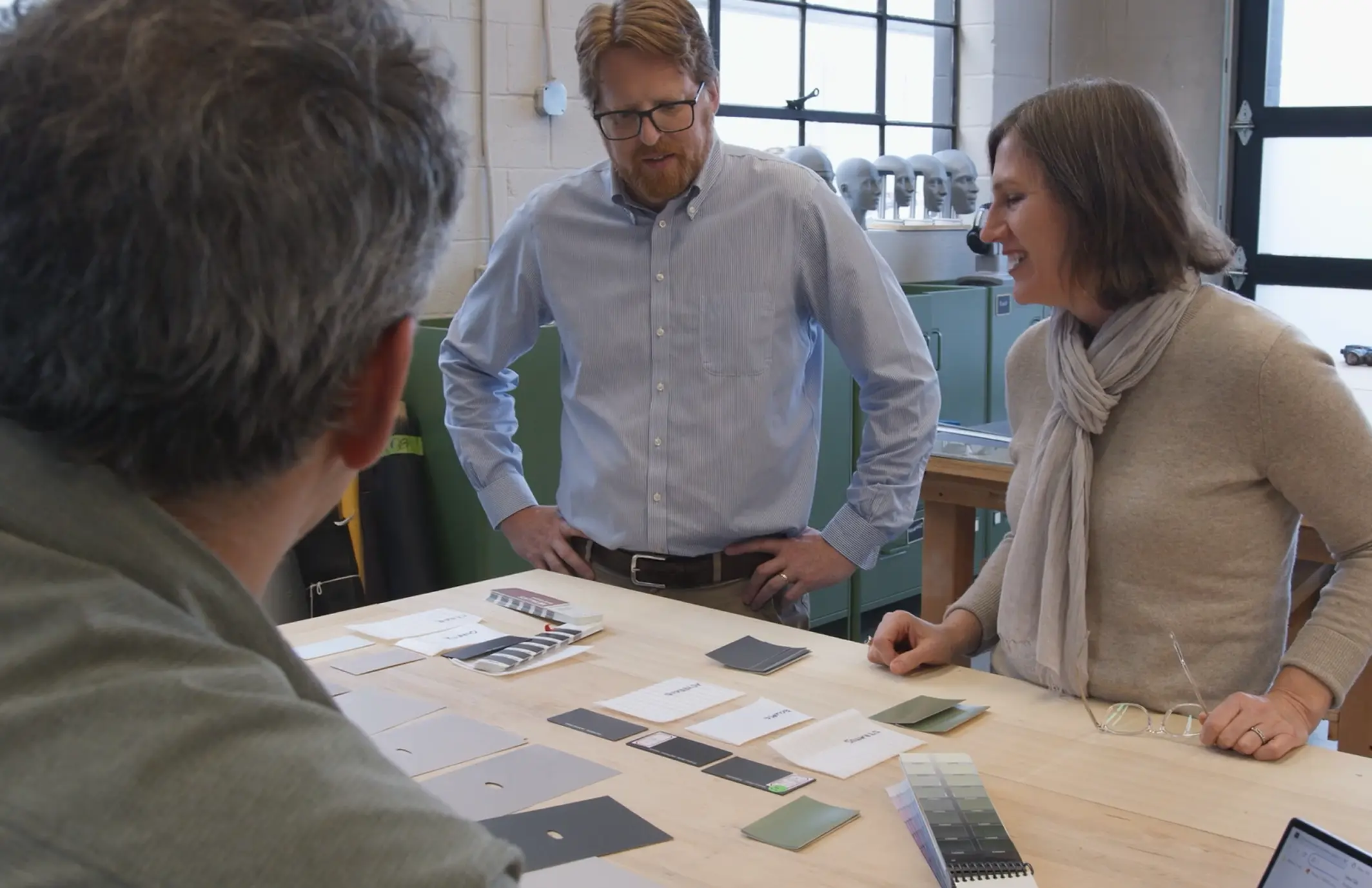 Three people standing around a wooden table with various paint samples and color swatches spread out, engaged in a discussion in a well-lit workshop or studio.