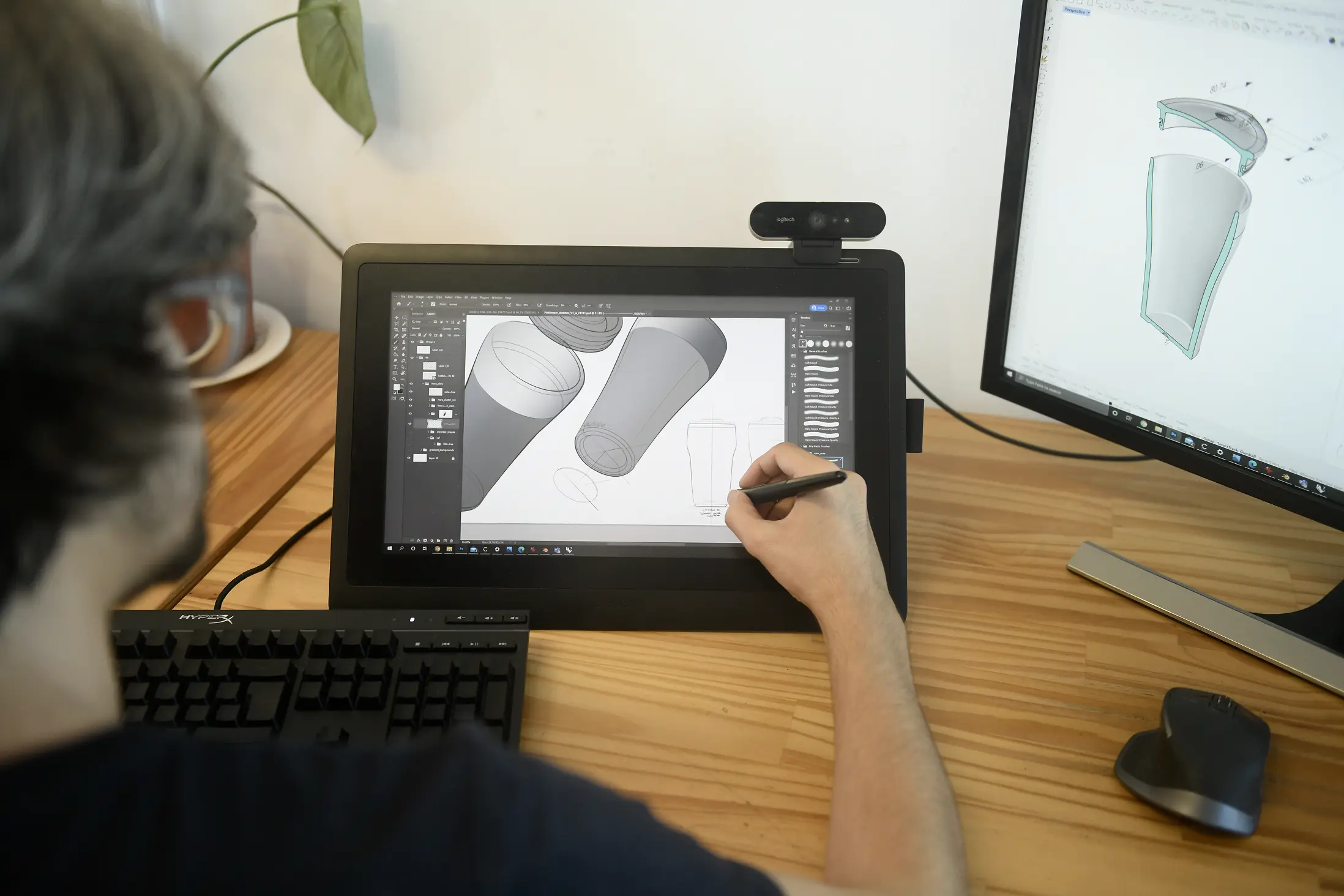 Person using a stylus on a drawing tablet displaying digital sketches of a cylindrical container, with a second monitor showing a 3D exploded view of the container's parts on a wooden desk.