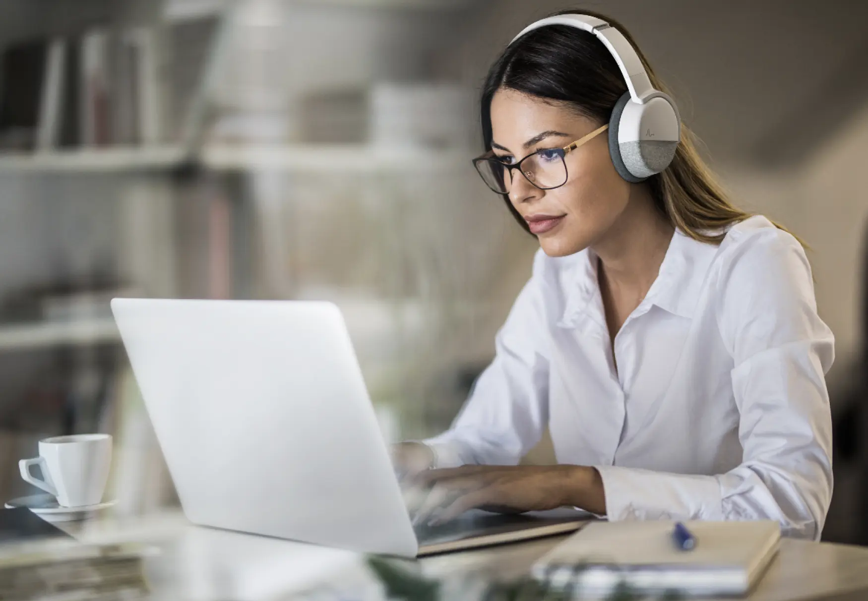 Woman wearing glasses and white headphones, focused on working on a laptop at a desk with a notebook and a cup of coffee nearby.