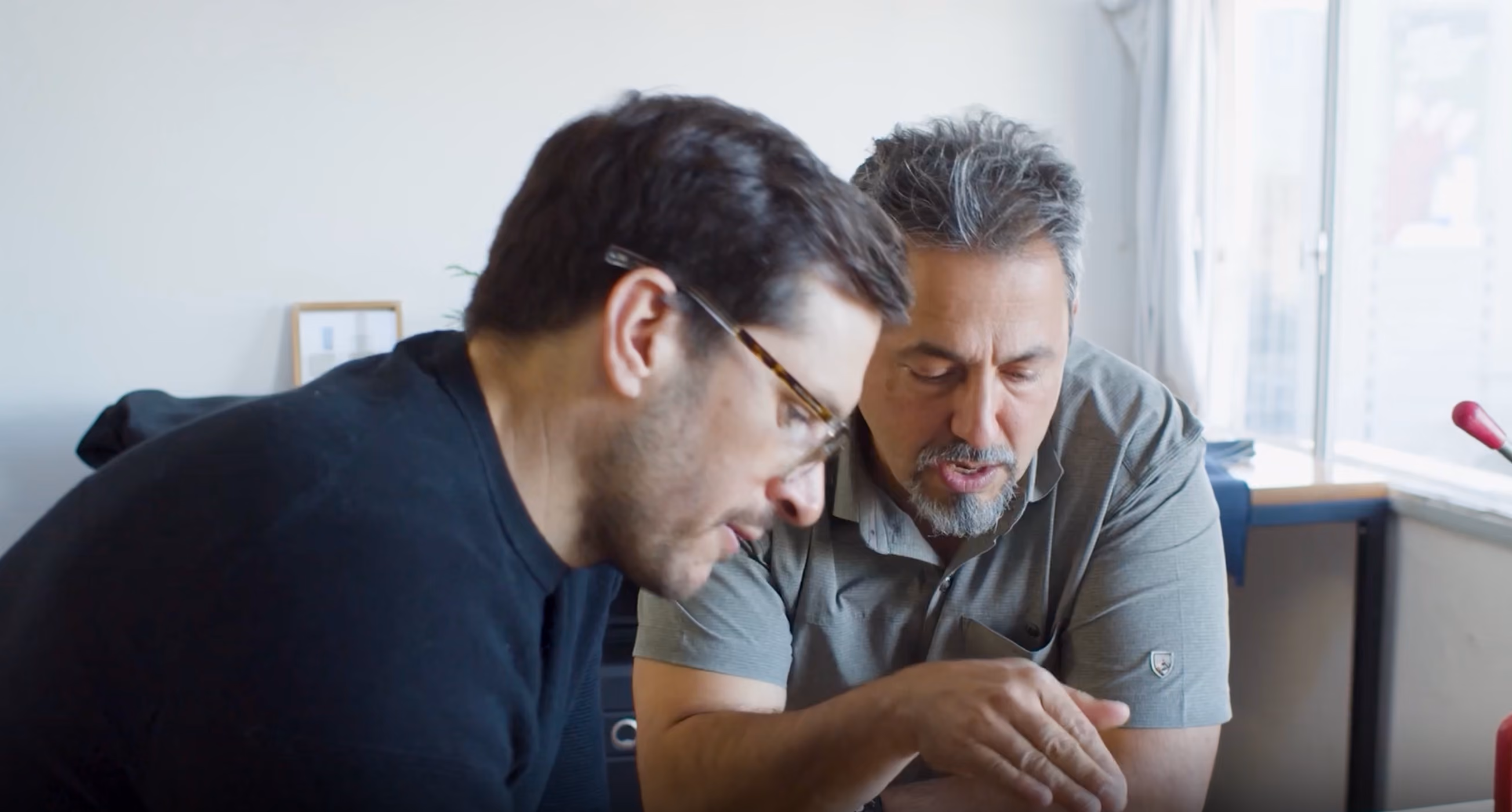 Two men sitting indoors, closely looking and discussing something with focused expressions, one man wearing glasses.