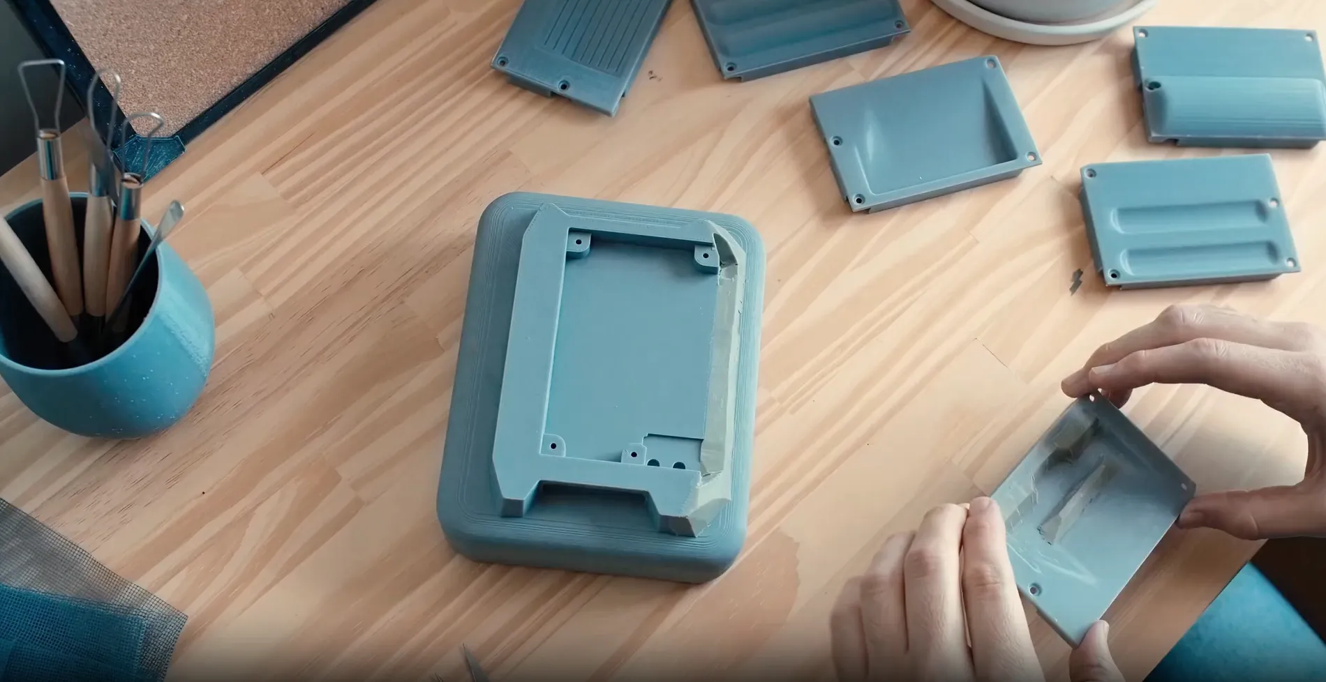 Two hands holding a small blue plastic mold piece over a matching base with other similar pieces on a light wooden surface, with a blue container holding various tools at the left edge.