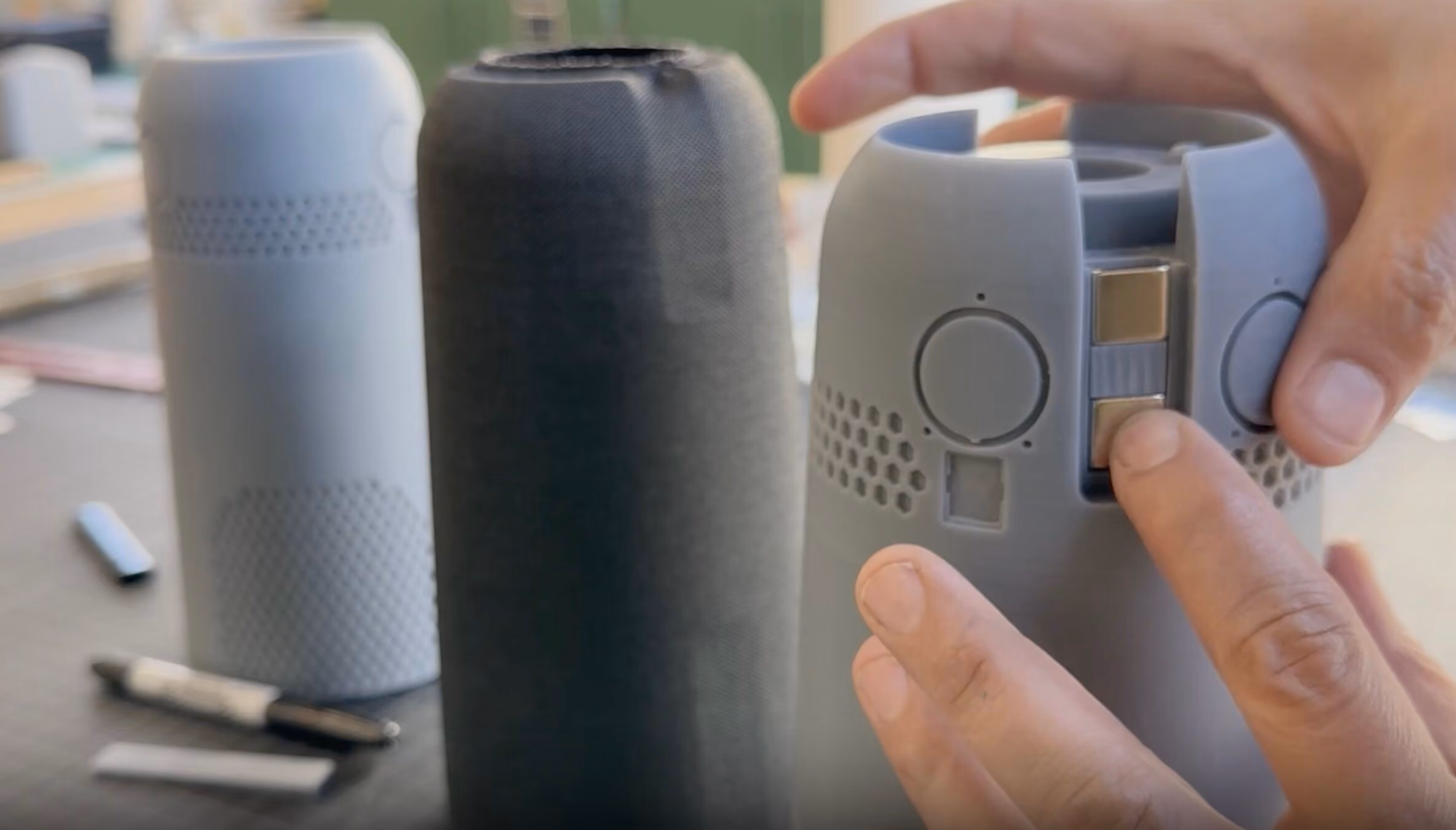 Close-up of hands assembling or adjusting a gray cylindrical smart speaker with round buttons and a sliding switch on the side, with two similar speakers in white and black blurred in the background on a table.