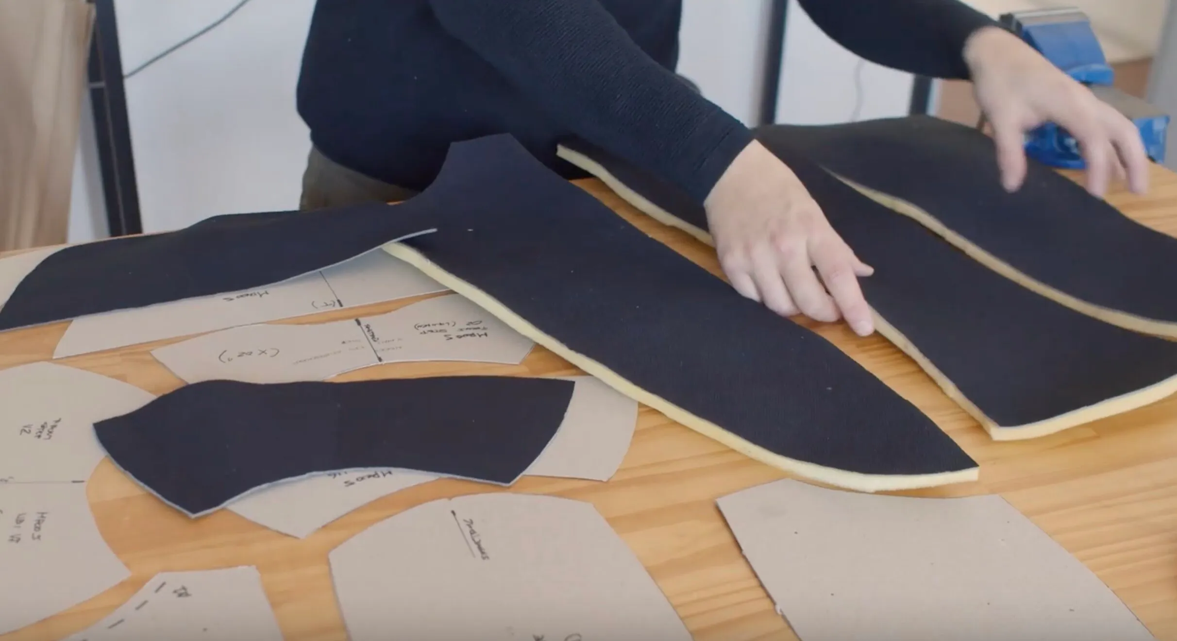 Person arranging black fabric pieces layered with yellow foam on a wooden table surrounded by paper sewing patterns.