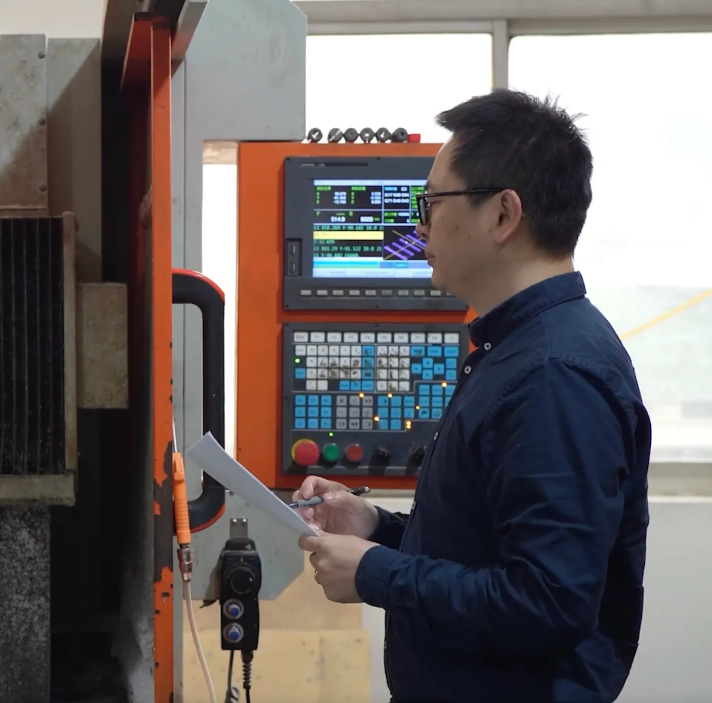 Man wearing glasses and a dark blue shirt holding a pen and paper while standing in front of an industrial machine with an orange control panel displaying data.