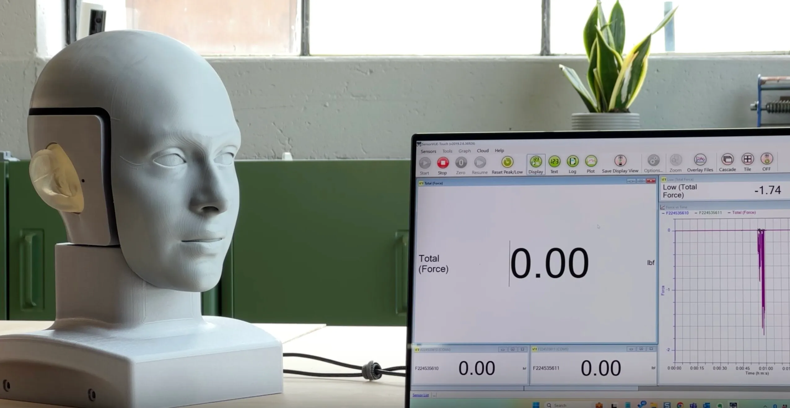 A white mannequin head with a realistic ear on the left side is placed on a table next to a computer monitor displaying a force measurement software interface showing zero total force and a graph with a purple line.