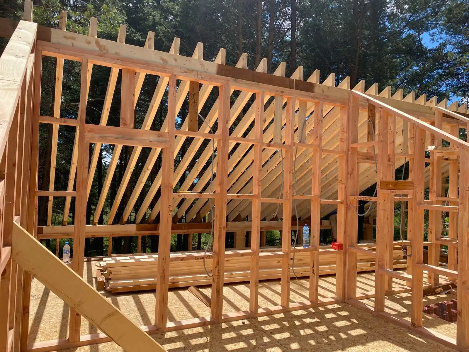 Wooden framework of a house under construction with exposed vertical and diagonal beams and roof rafters, set outdoors with trees in the background.