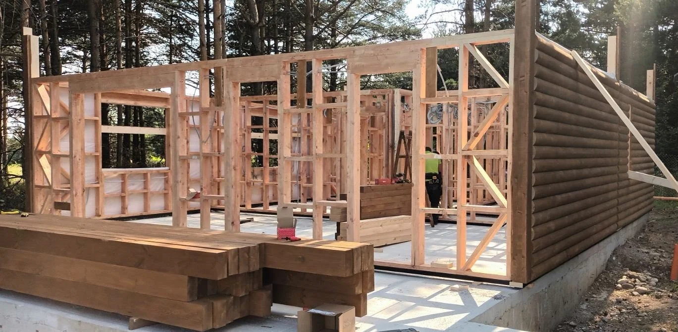 Wooden frame structure of a building under construction on a concrete foundation, with stacked wooden beams in front and surrounded by trees.