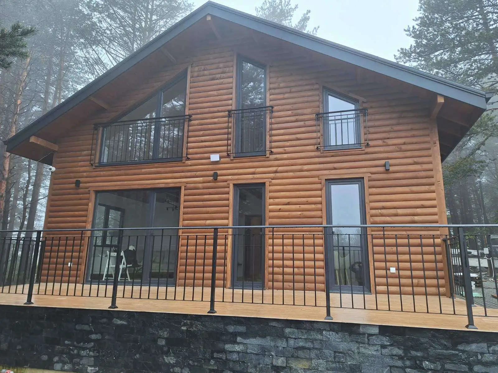 Two-story wooden cabin with log-style siding and large black-framed windows and doors, featuring a balcony and deck with black metal railing, set against a foggy forest background.