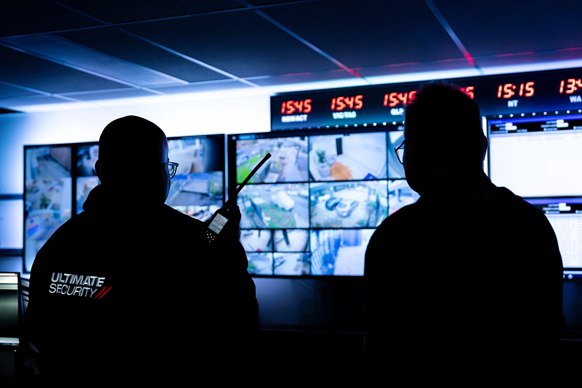 Two Ultimate Security staff silhouetted in a dark monitoring room, watching a wall of live CCTV camera feeds.