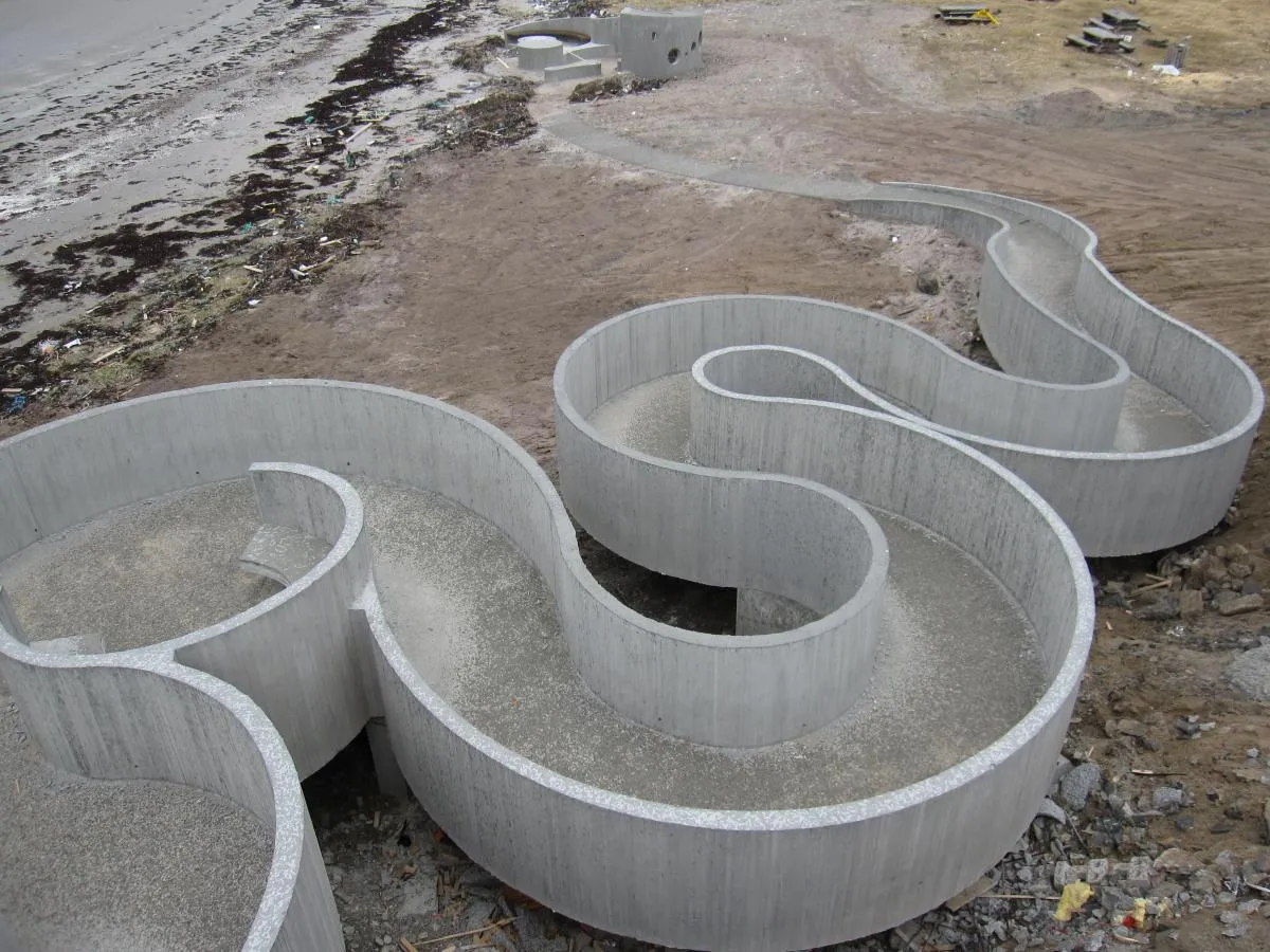 To store slangeformede betongkonstruksjoner på bakken nær en strand med tang og sand.