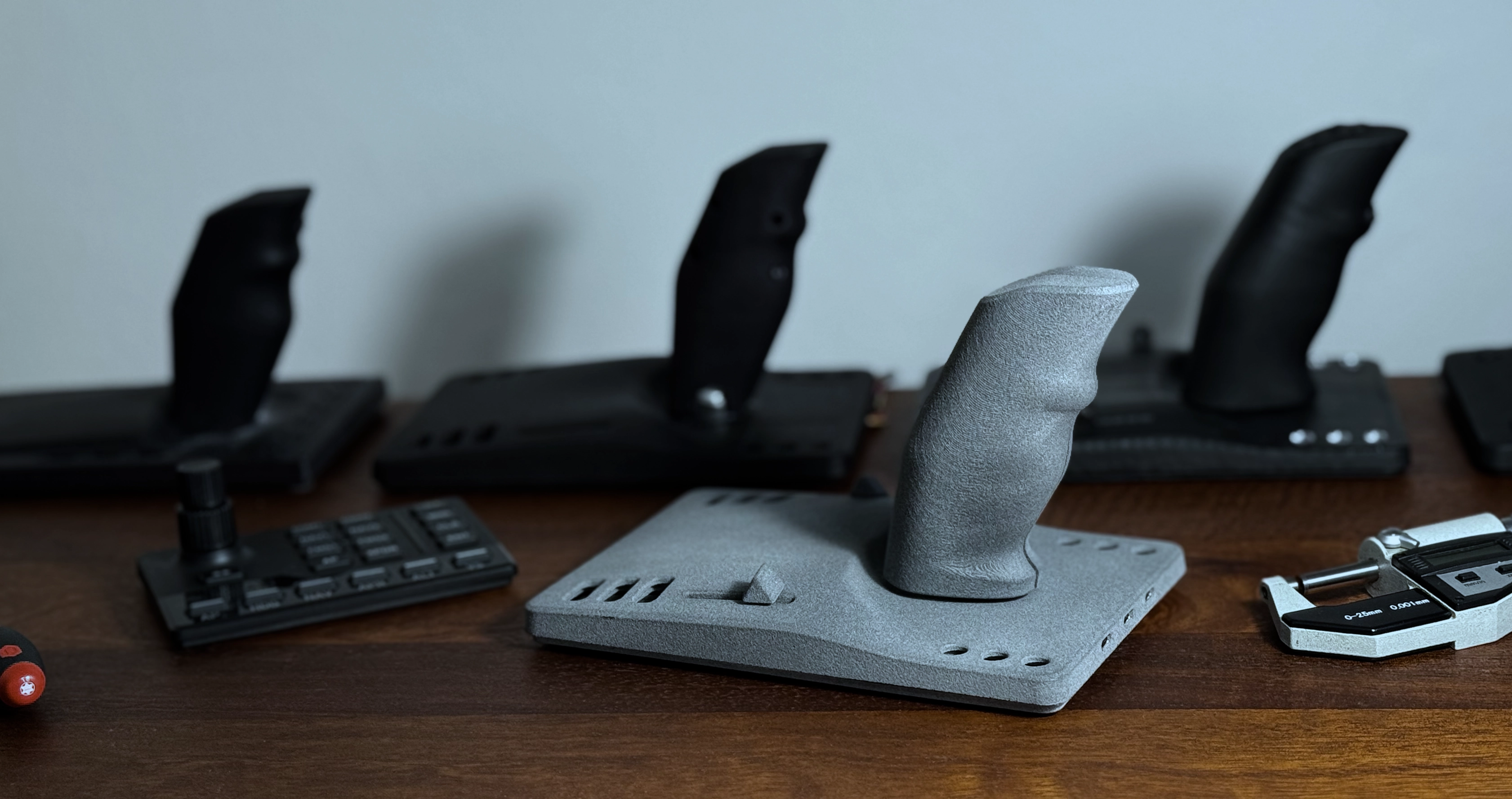 A textured gray joystick on a base placed on a wooden surface, surrounded by other black joysticks and a measuring tool.