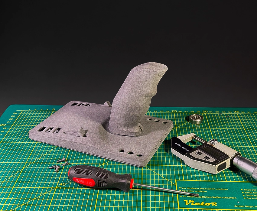 Orange and gray 3D-printed mechanical parts placed on top of a technical drawing, with a digital caliper nearby and 3D printers blurred in the background.