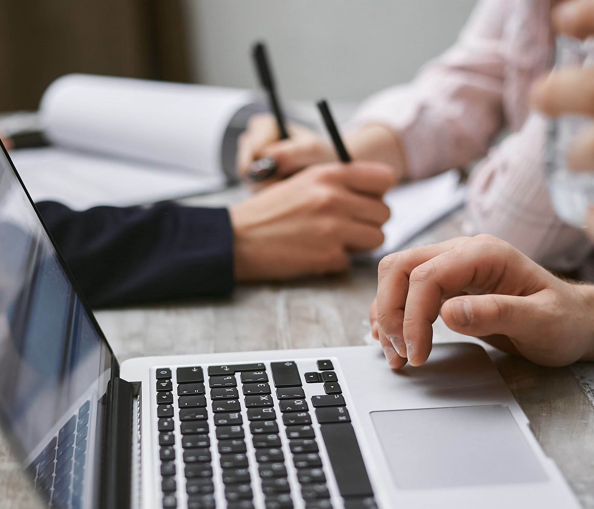 Close-up of hands at a wooden table with a laptop keyboard and two people writing in notebooks with black pens.