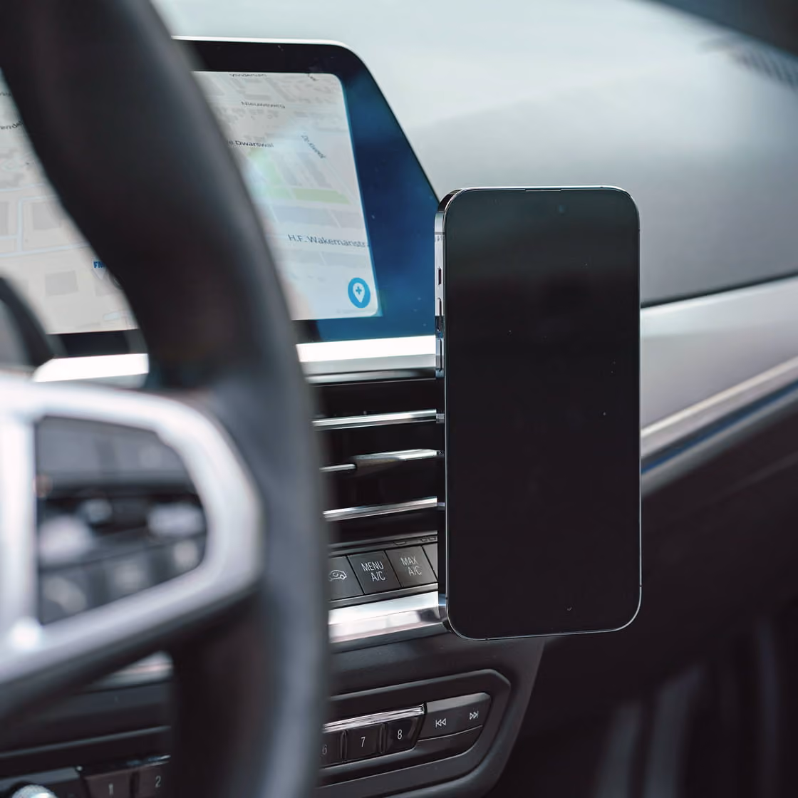 Inside a car, a smartphone with a black screen is mounted on an air vent near the dashboard, next to a digital navigation display and partially visible steering wheel.