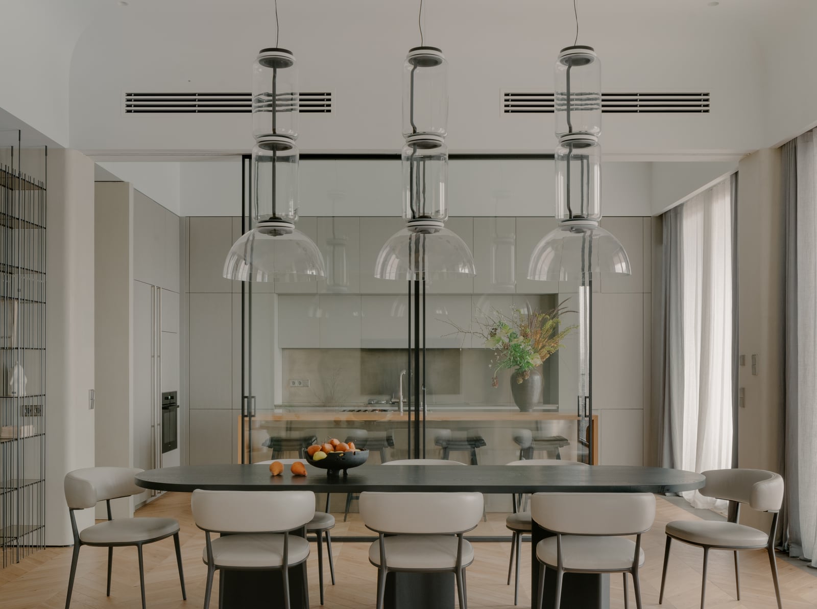 Dining room with an oval table and six chairs under three glass pendant lights, kitchen behind a glazed partition