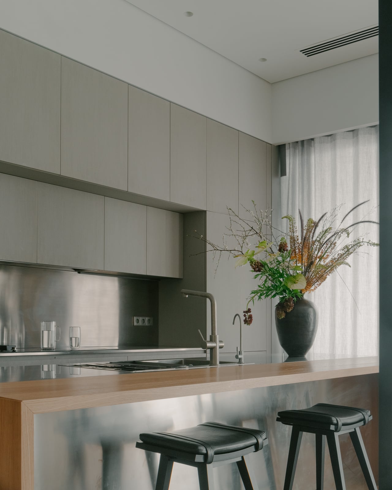 Kitchen island with a wooden counter, black stools and a stainless steel splashback, flowers in a vase