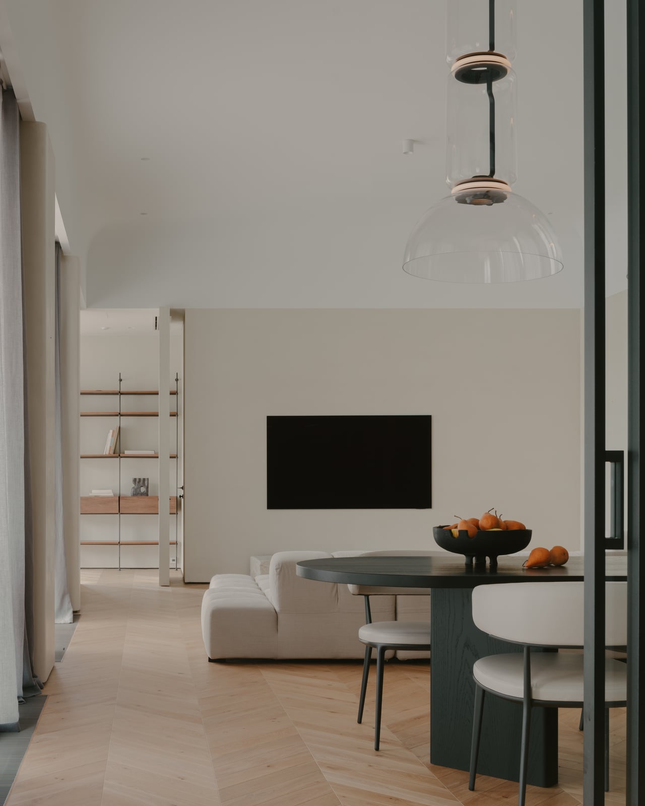 Living room seen past open shelving: sofa, round dining table and a wall-mounted screen