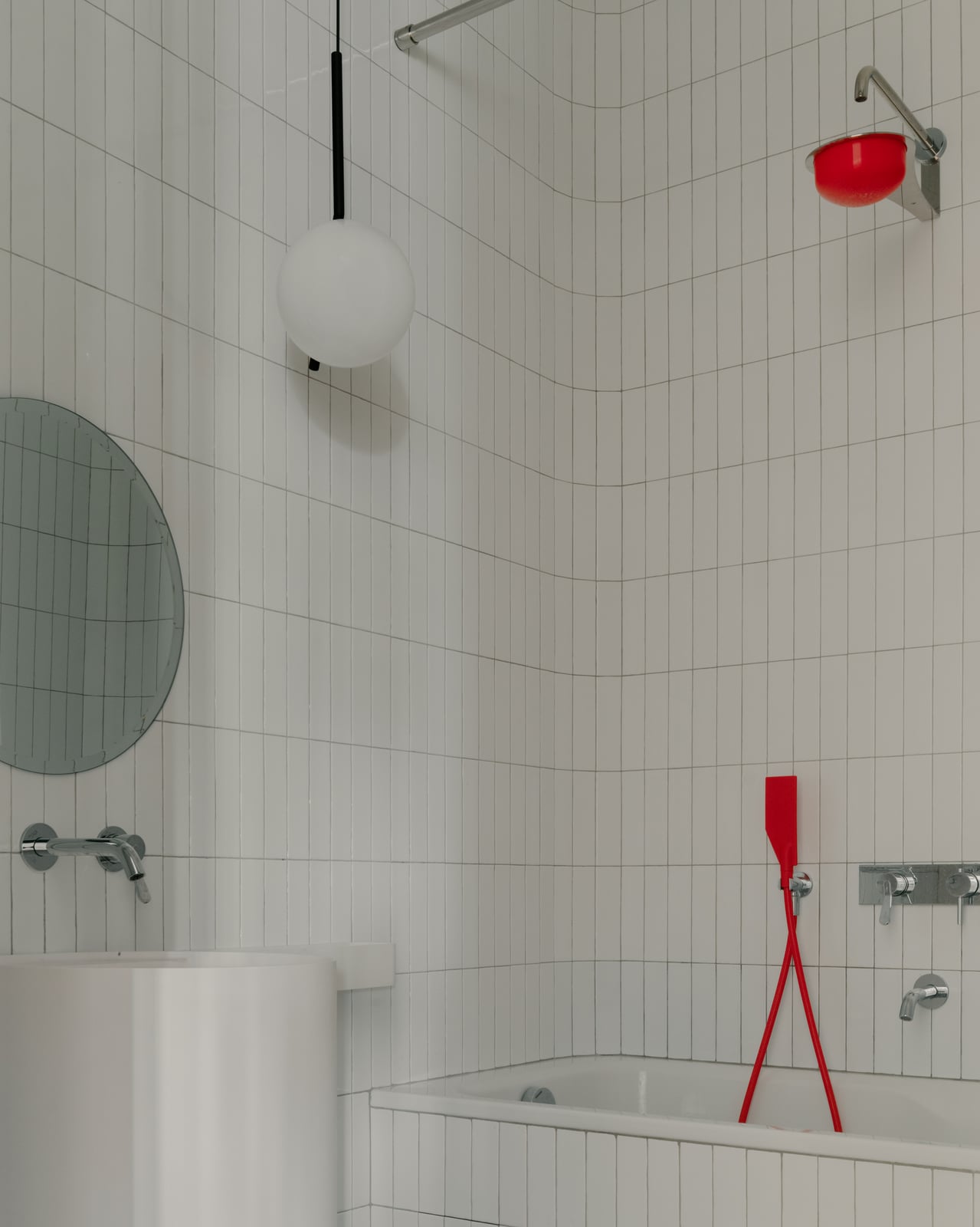White tiled bathroom with a round mirror, a globe light and red fittings