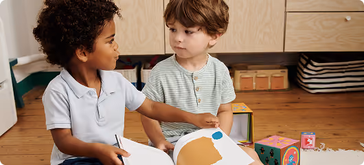 Deux jeunes garçons assis sur le sol en bois, l'un montrant un livre ouvert à l'autre, entourés de jouets colorés.