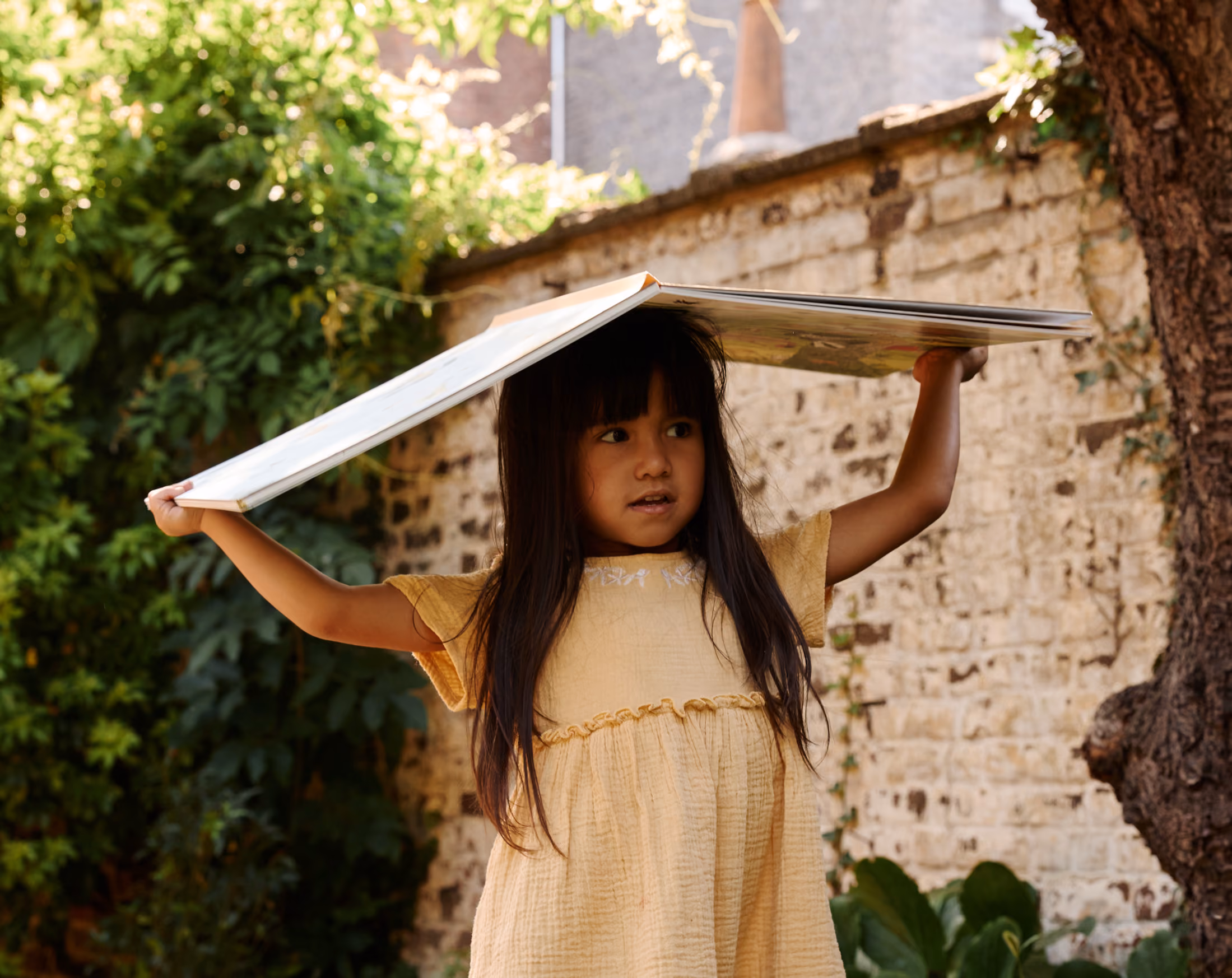 Petite fille aux longs cheveux noirs portant une robe jaune claire tenant un grand livre ouvert au-dessus de sa tête à l'extérieur près d'un mur en pierre et un arbre.