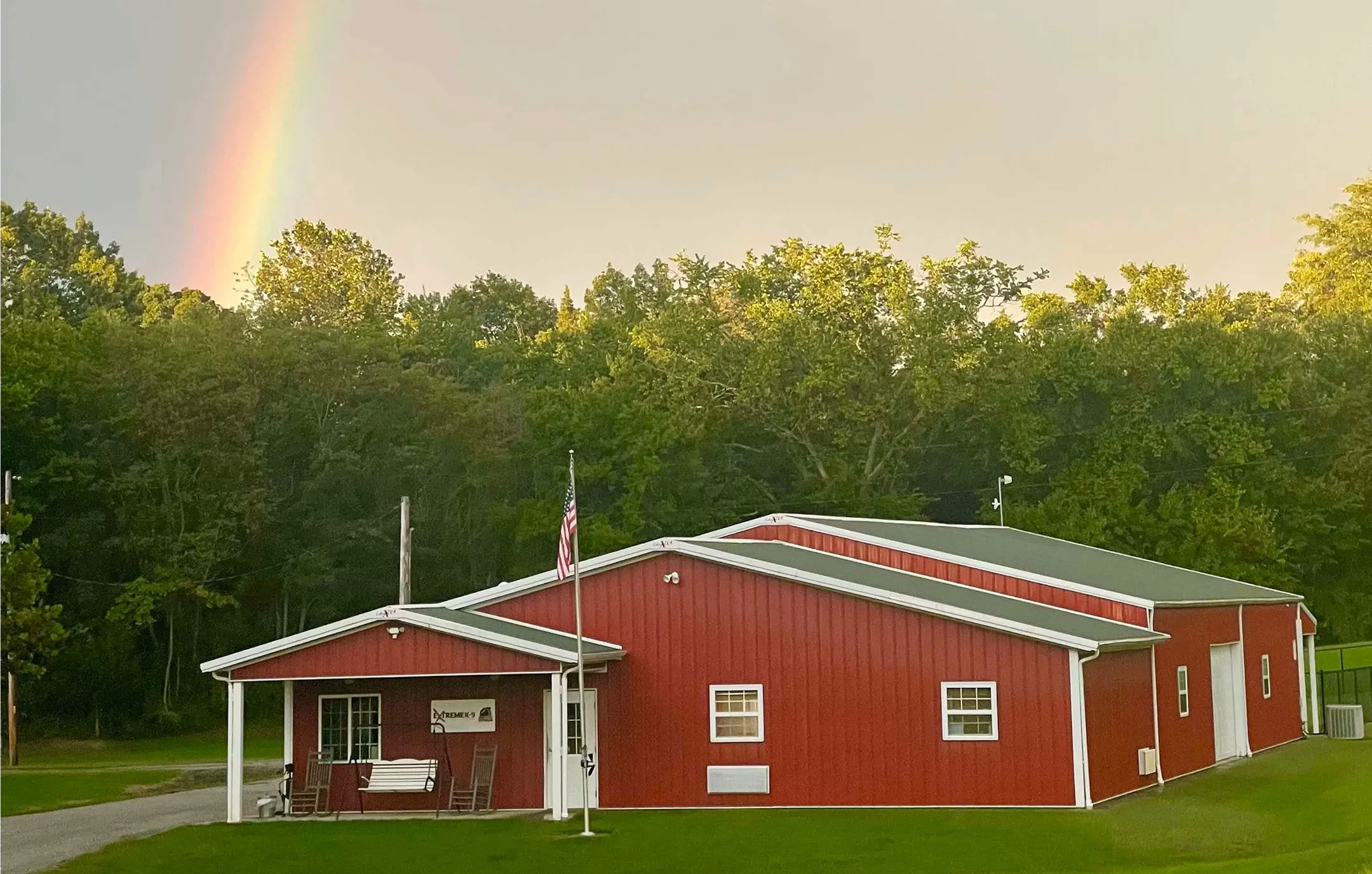 A large red barn-style building with a covered front porch featuring a white bench and rocking chairs, an American flag on a pole in front, surrounded by green grass and dense trees with a rainbow visible in the sky to the left.