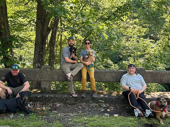 Three people outdoors on a sunny day sitting and standing on a large stone bench surrounded by trees, each accompanied by a dog; one man on the left crouches beside a black dog, a man and woman in the center hold smaller dogs while sitting on the bench, and a woman on the right sits on the ground with a large dog on a leash.