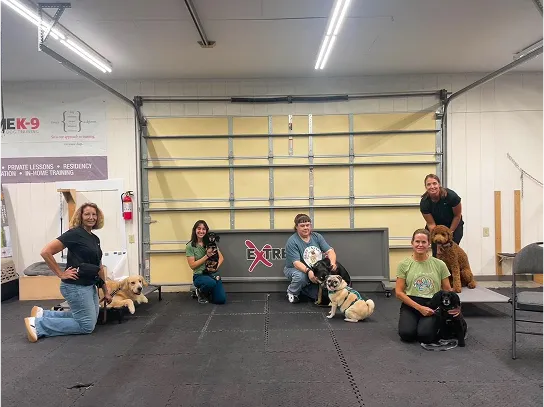Four women kneeling on a black floor indoors, each posing with a dog of various breeds in front of a large beige garage door, with an 'Extreme K-9' sign visible behind them.