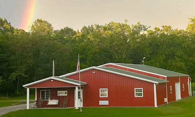 Red building with a white roof and flagpole displaying an American flag, surrounded by green grass and trees with a rainbow visible in the sky.