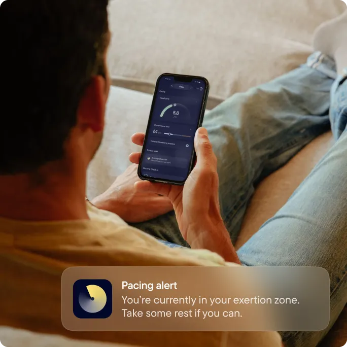 A man relaxing on a sofa while checking the Visible app on his phone. A notification reads ‘Pacing alert: You’re currently in your exertion zone. Take some rest if you can.’