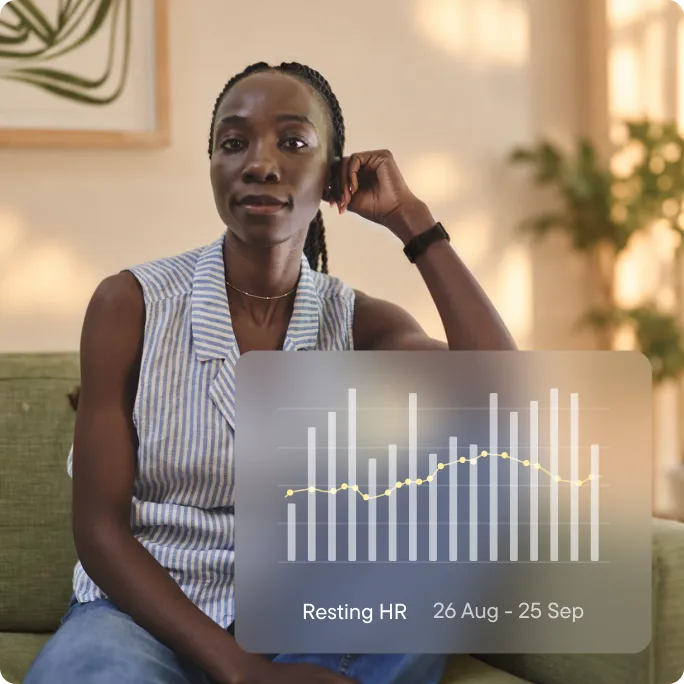A woman sitting on a sofa at home, wearing a Visible band on her wrist. An overlay shows a bar chart of her resting heart rate from 26 August to 25 September.