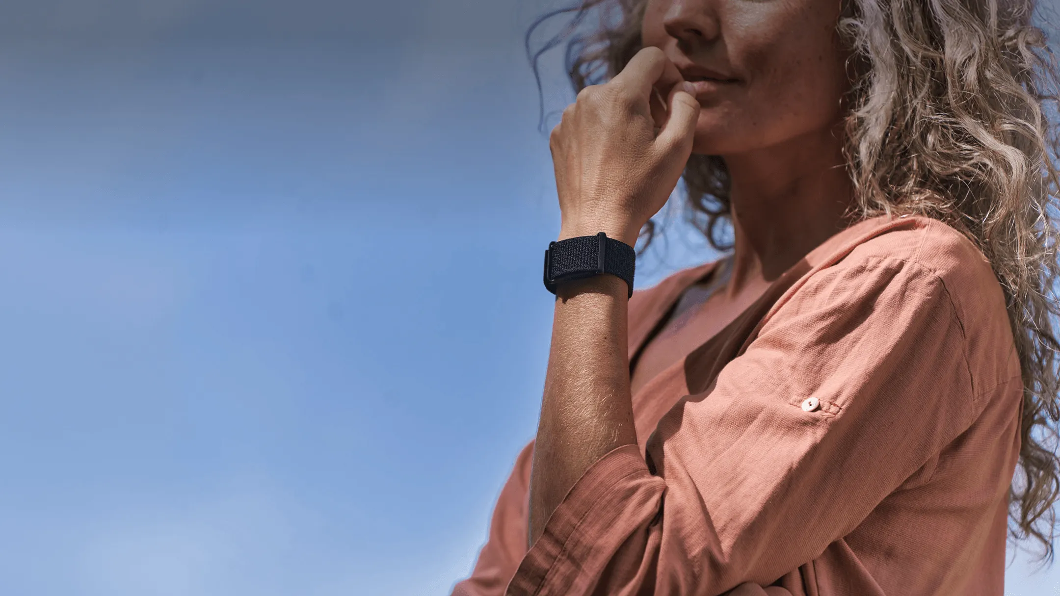 A woman in a coral linen shirt rests her hand near her chin, wearing a black Visible band on her wrist.