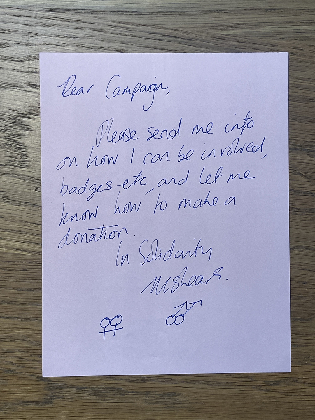 Handwritten note on white paper placed on a wooden surface, reading: 'Dear Campaign, Please send me info on how I can be involved, badges etc, and let me know how to make a donation. In Solidarity, Ms Spears.' with two pairs of interlinked gender symbols at the bottom.